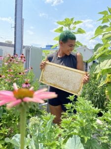 Lead Harvester, Che holds a beehive in a lush garden.