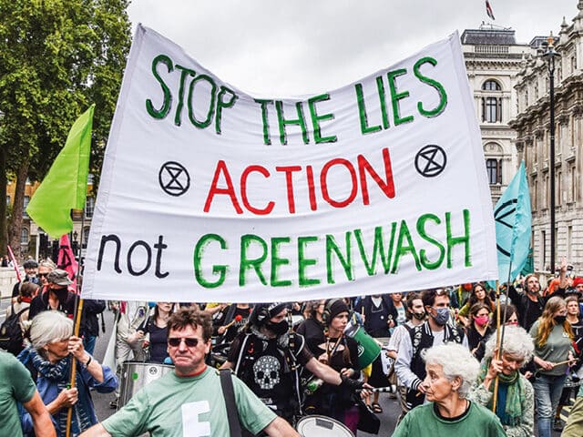 Protests march in the streets against greenwashing, holding a sign that says "stop the lies; action, not greenwash"