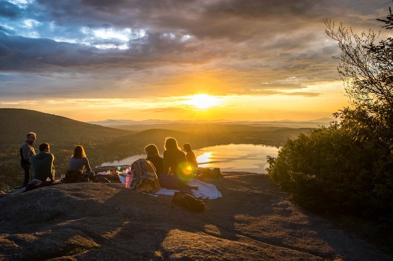 A group of people sitting and looking out over a lake and distant mountains at sunset